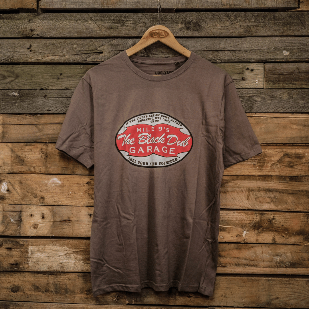 A gray crew neck t-shirt with 'The Black Dub Garage' text printed on the front, displayed against a wooden backdrop.