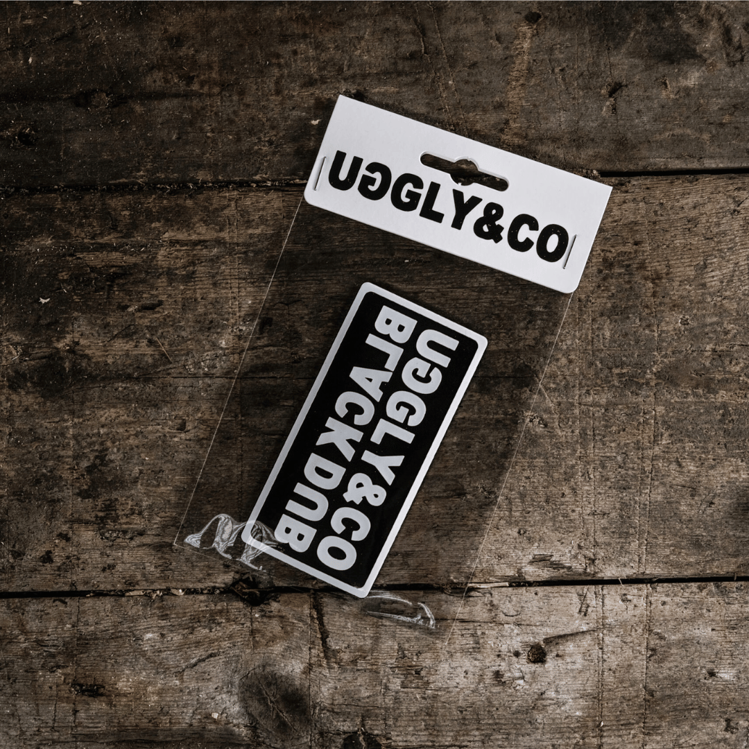 A black and white patch with text, placed on a wooden surface, with the brand name 'UGLY&CO' visible above it.